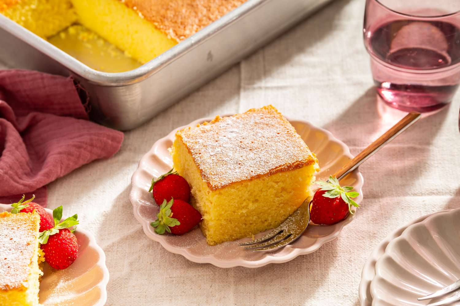 A slice of cake on a plate with strawberries and a fork set on a table with another slice and a baking dish in the background