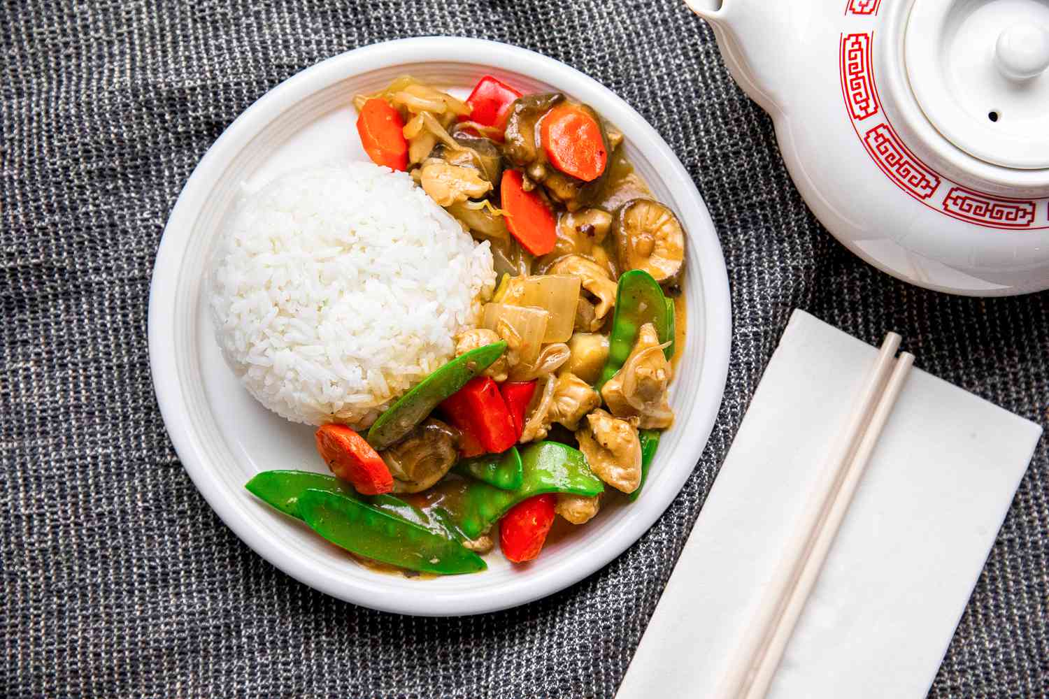 Plate of Chop Suey With Rice Next to a Pair of Chopsticks on a Napkin and a Teapot, All on a Grey Kitchen Towel
