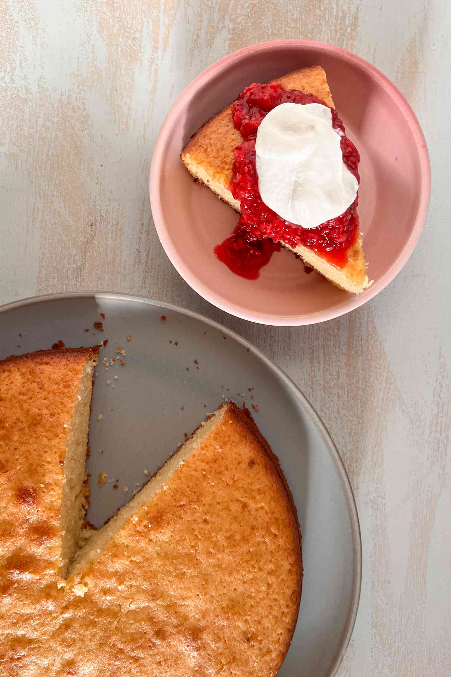 Overhead shot: slice of French yogurt cake on a plate with raspberry sauce and whipped cream and more cake on a plate next to it