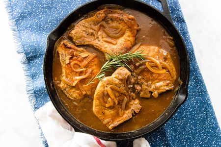 Four gluten free Smothered Pork Chops with Gravy in a skillet with a sprig of rosemary on top set against a blue background.
