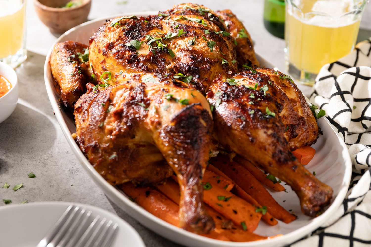 Smoked Spatchcock Chicken with Harissa Butter and Grilled Carrots in a Bowl, Surrounded by Drinks, Kitchen Towel, and Bowl with Two Forks