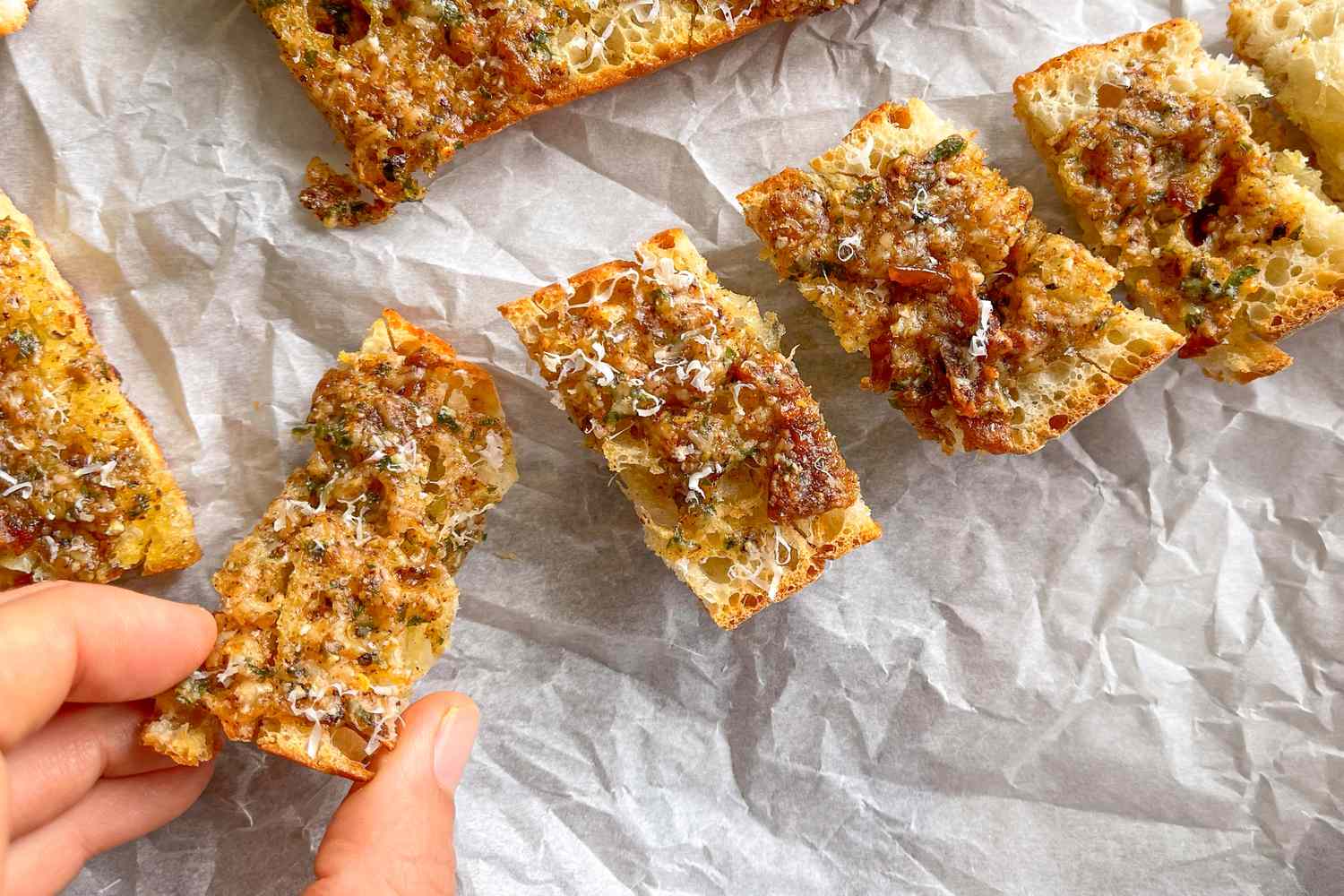 Garlic bread on a piece of wrinkled parchment paper; a hand coming into frame to grab a piece