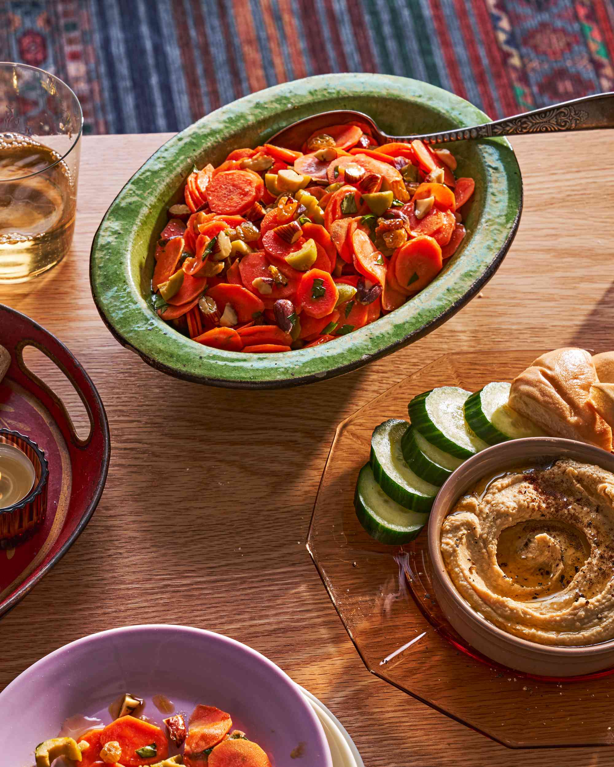 A bowl of carrot salad on a coffee table, alongside hummus with cucumbers and pita and a glass of white wine