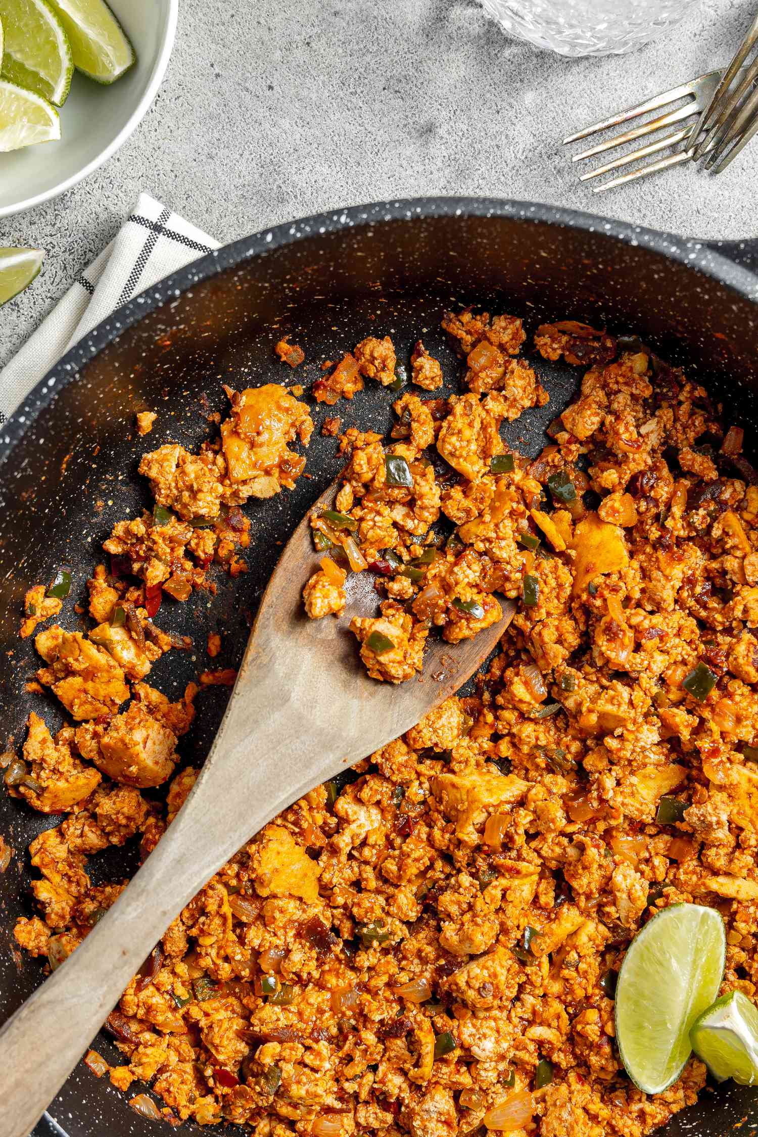 Copycat Sofritas in a Pan with a Wooden Spoon and Lime Wedges, Surrounded by a Bowl of Lime Wedges, a Glass of Water, and a Couple of Forks