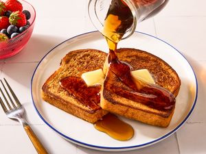 French toast with butter and syrup being poured accompanied by a bowl of berries and a fork on the side