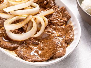 Plate of Bistek Topped With Onions Next to a Bowl of Rice