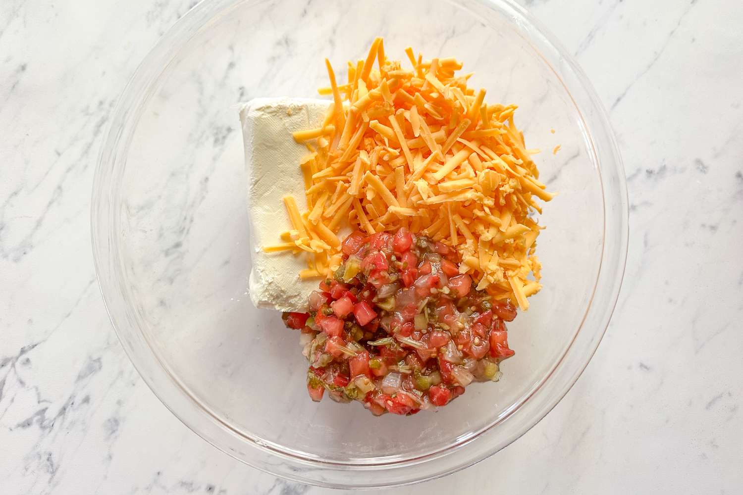 Ingredients for a dip in a bowl including cream cheese shredded cheese and diced tomatoes with other seasonings