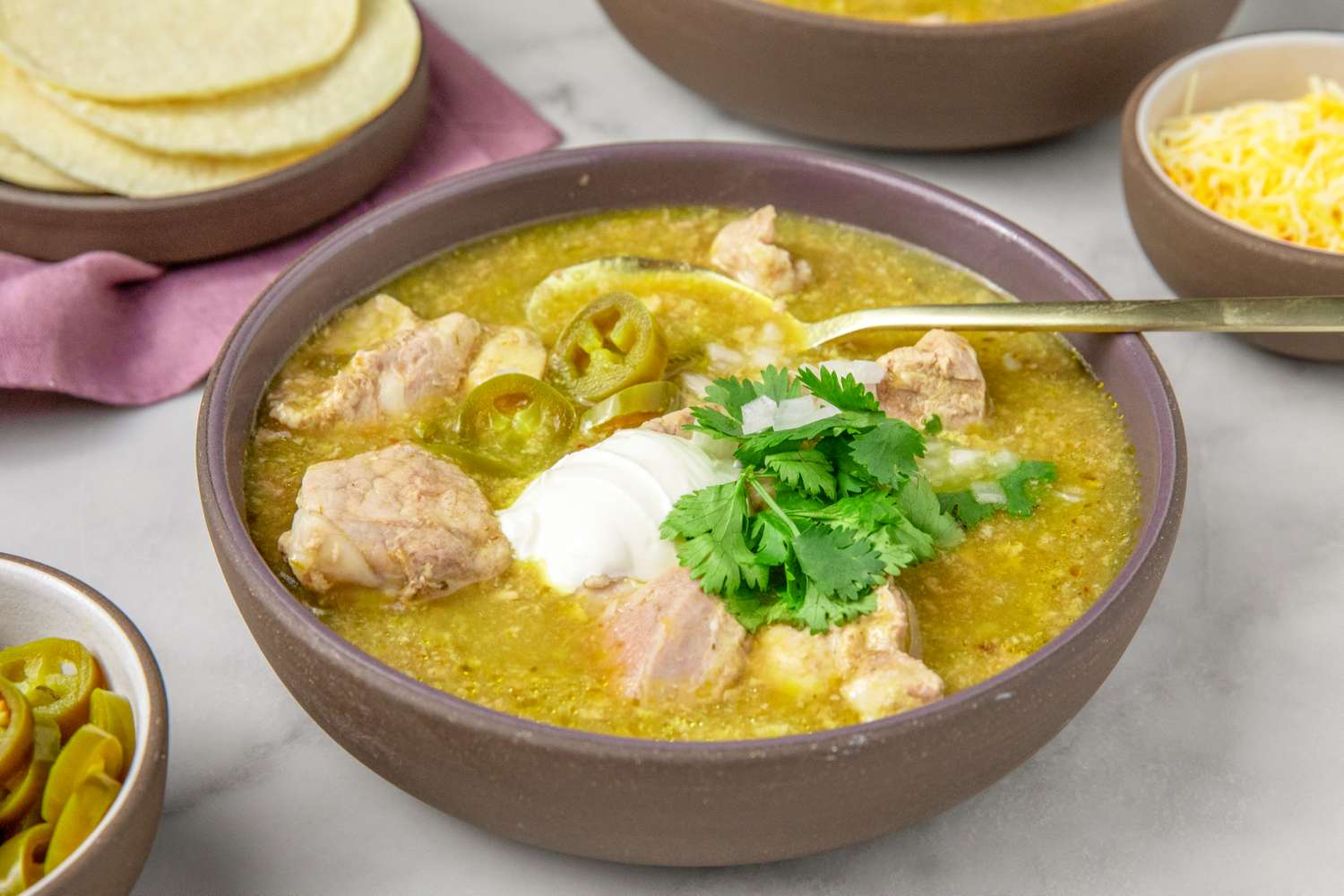 A bowl of pork chile verde garnished with sour cream cilantro and jalapeo slices accompanied by tortillas and cheese on the side