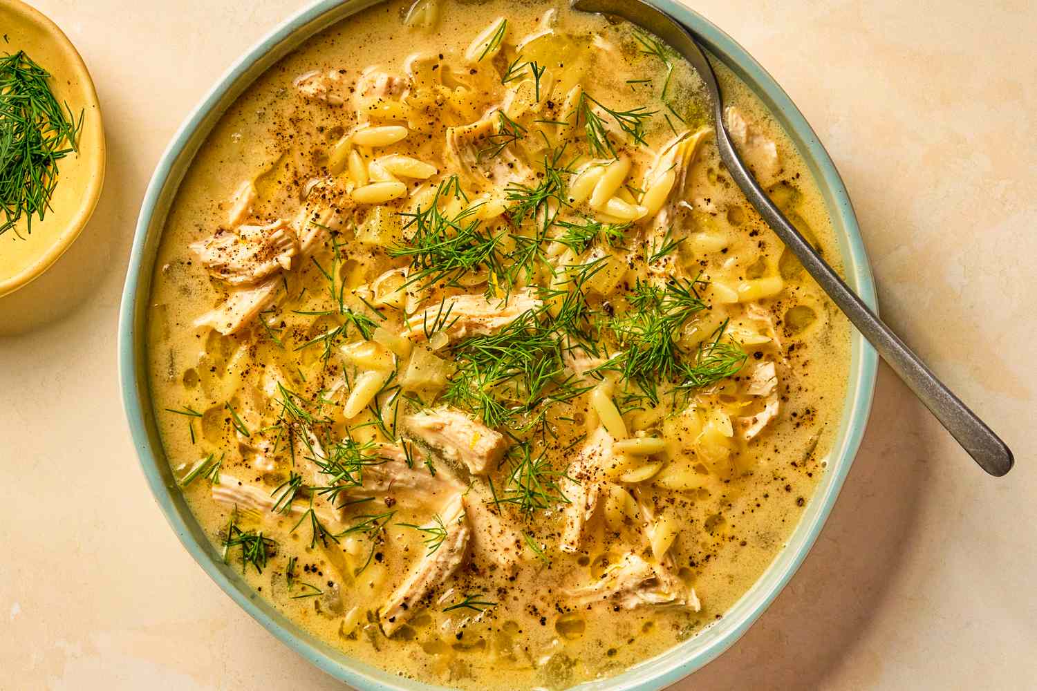 A bowl of soup with orzo pasta shredded chicken and fresh dill garnish spoon resting on the edge