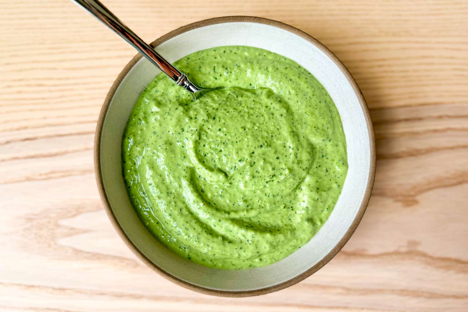 Creamy green dip in a bowl with a spoon, served on a wooden surface.