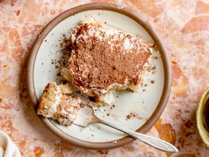 Plate with a serving of tiramisu topped with cocoa powder and a fork placed on the side small piece removed from the dessert