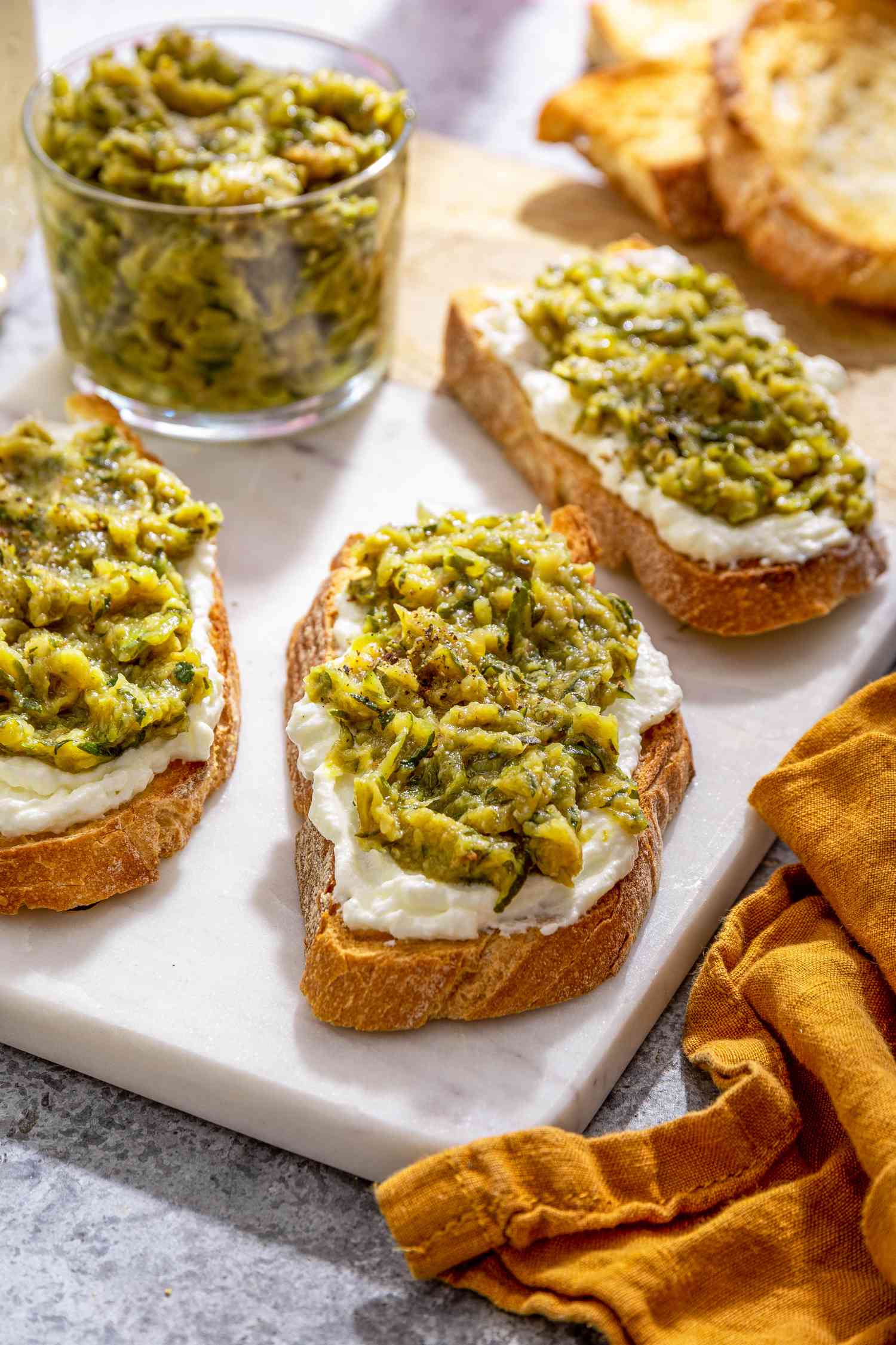 Slices of Toast Topped With Ricotta and Zucchini Butter on a Marble Slab Next to a Bowl With More Zucchini Butter and a Spoon, and in the Surroundings, a Mustard Colored Kitchen Towel and a Glass