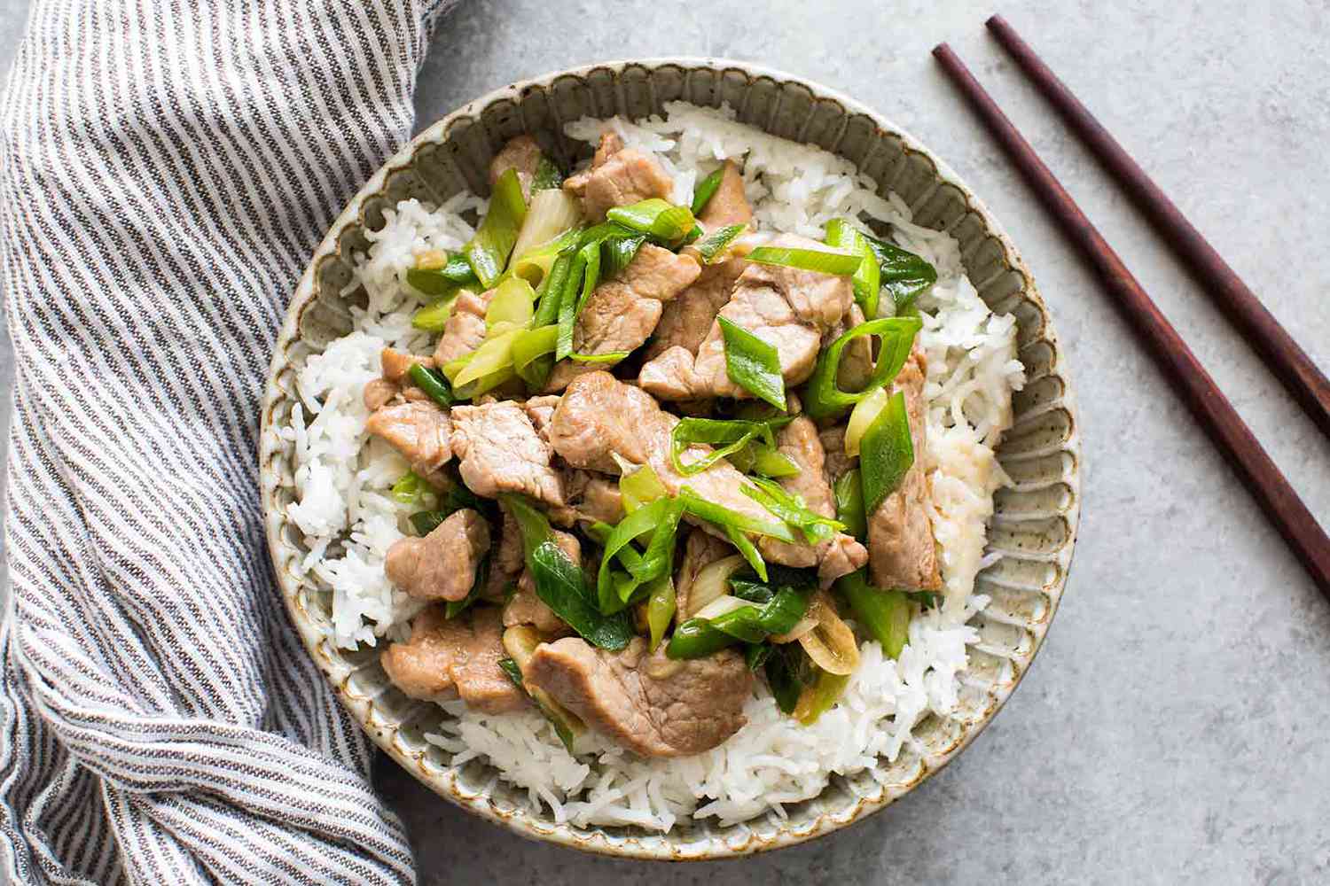 Bowl of pork stir fry with green onions served over rice alongside a striped cloth and chopsticks
