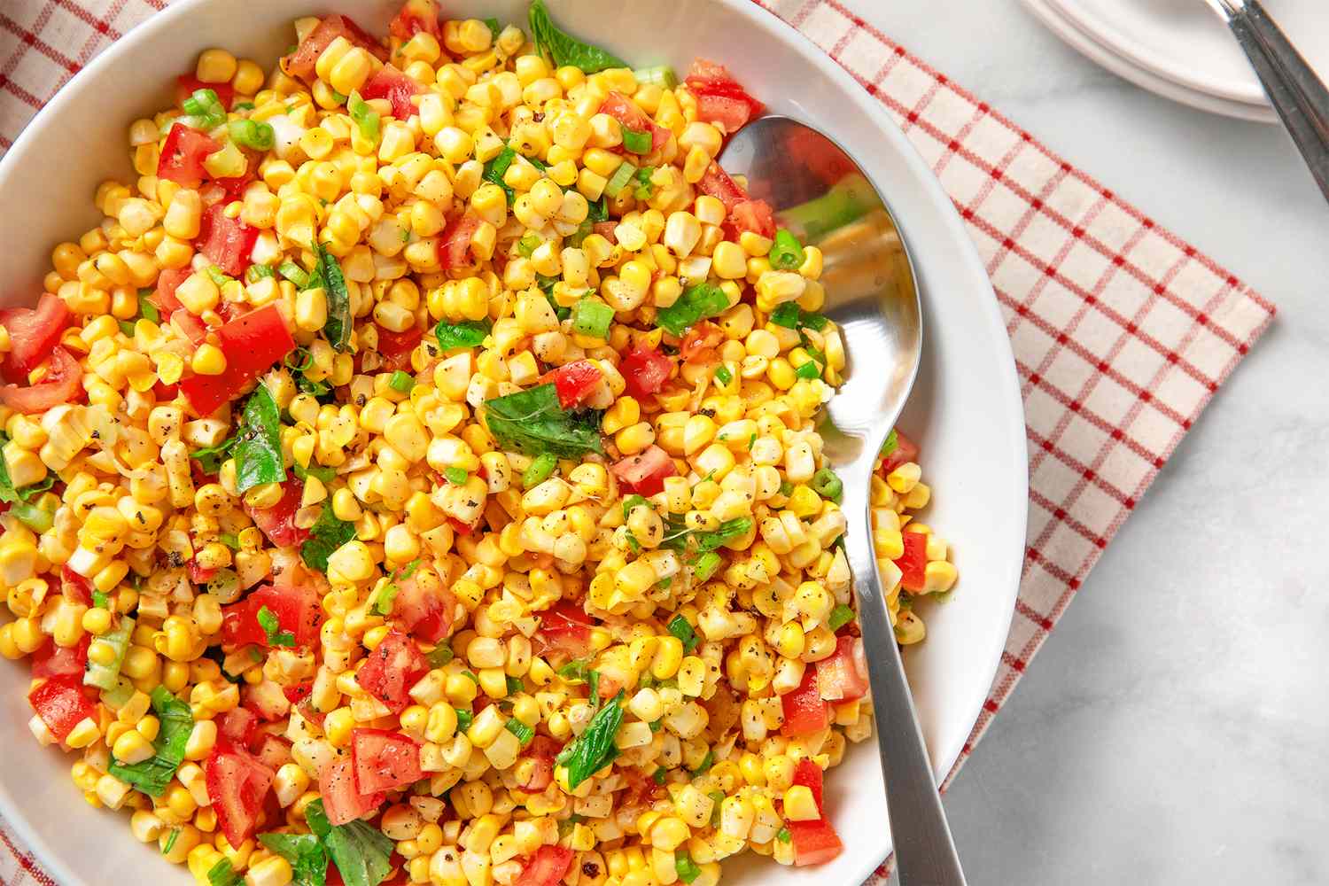 Bowl of corn salad with tomatoes herbs and a serving spoon positioned on a patterned napkin