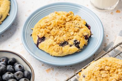 Blueberry Muffin Top on a Plate, Surrounded by a Bowl of Blueberries, More Muffin Tops on a Wire Rack, a Glass of Milk, and Another Muffin Top on a Plate