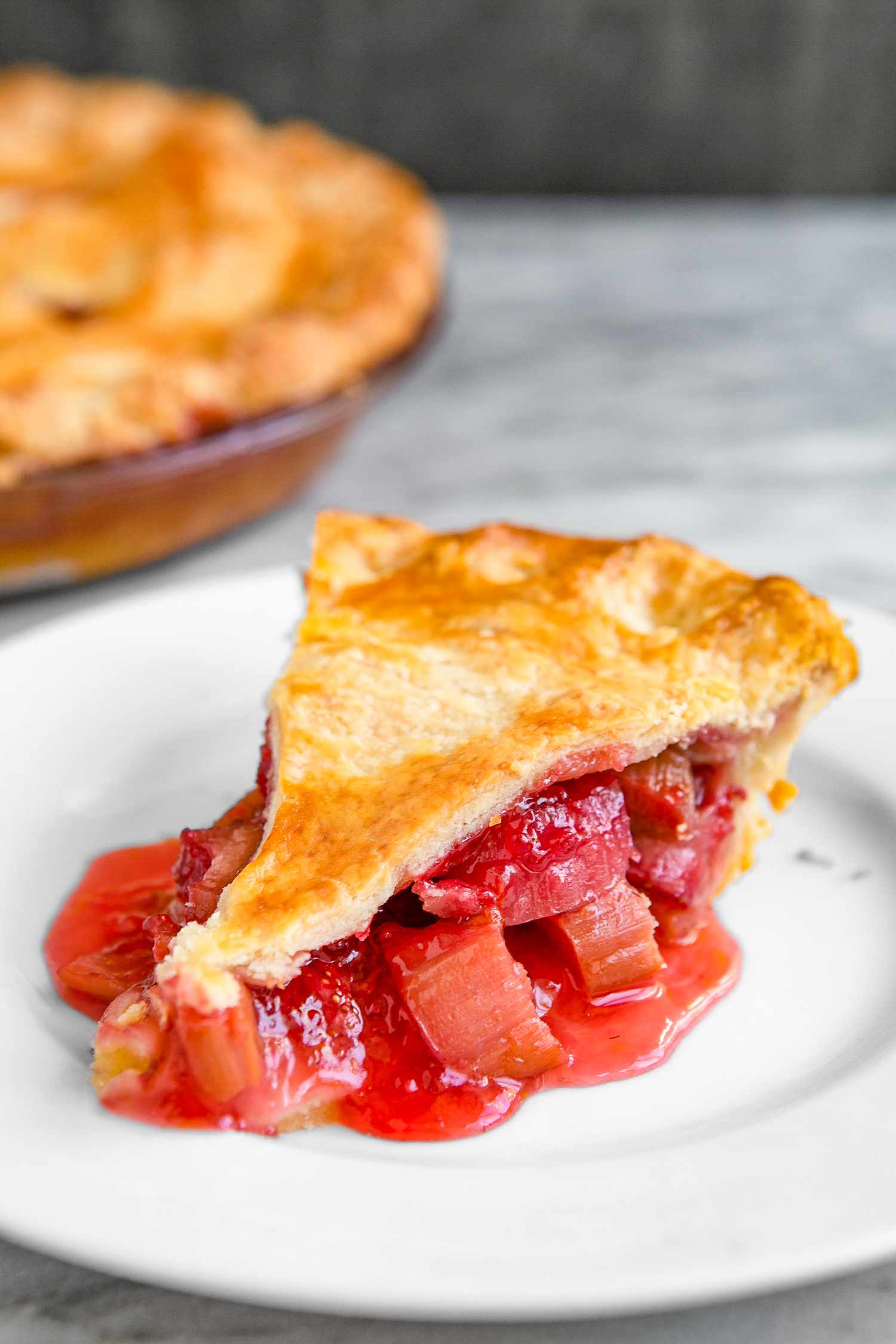 Slice of Strawberry Rhubarb Pie on a Plate, and in the Background, More in a Glass Pie Pan