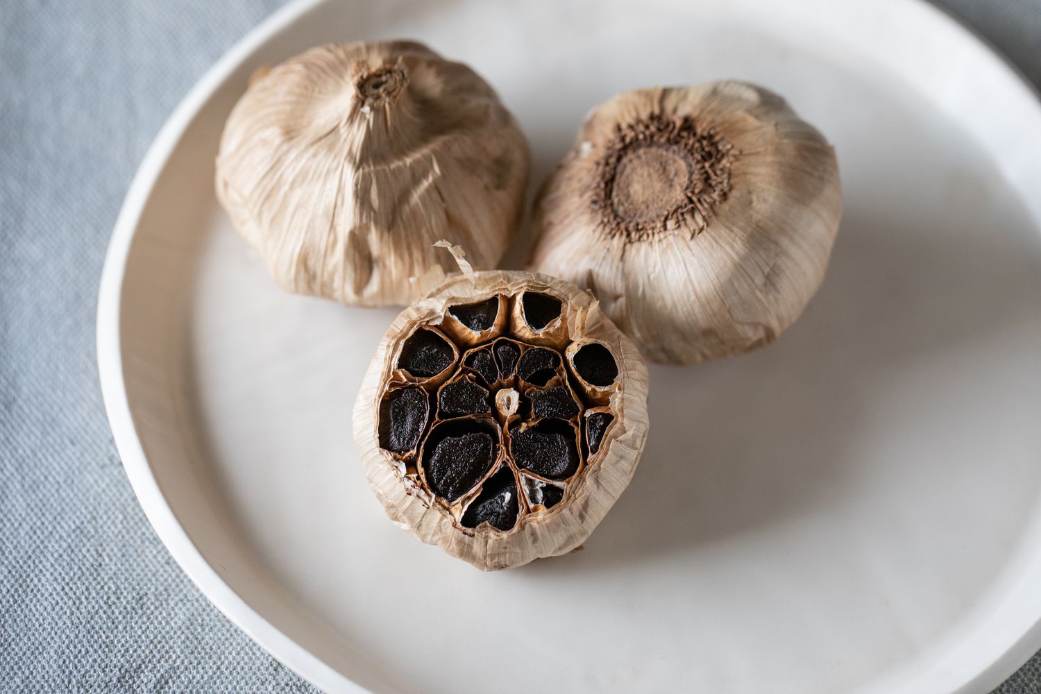 Three heads of black garlic on a plate with one of them sliced to show the cloves.