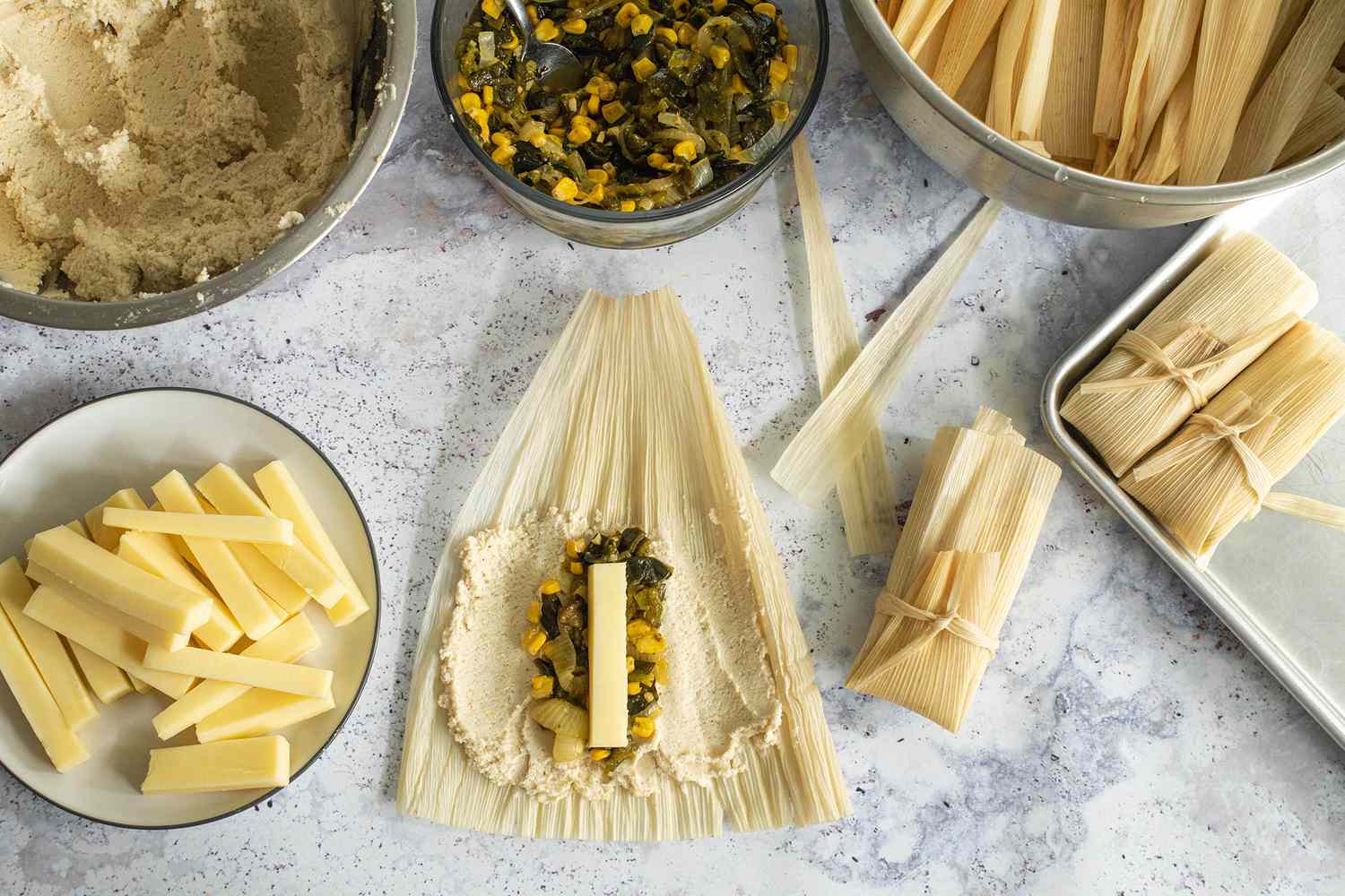 Making the Tamales: Corn Husk Covered in Masa and Topped with Poblano, Corn, and Cheese, and in the Surroundings, Plate of Cheese Strips, a Bowl of Masa, a Bowl of Pepper and Corn Filling, a Bowl of Corn Husks, and a Baking Tray with Poblano, Corn, and Cheese Tamales