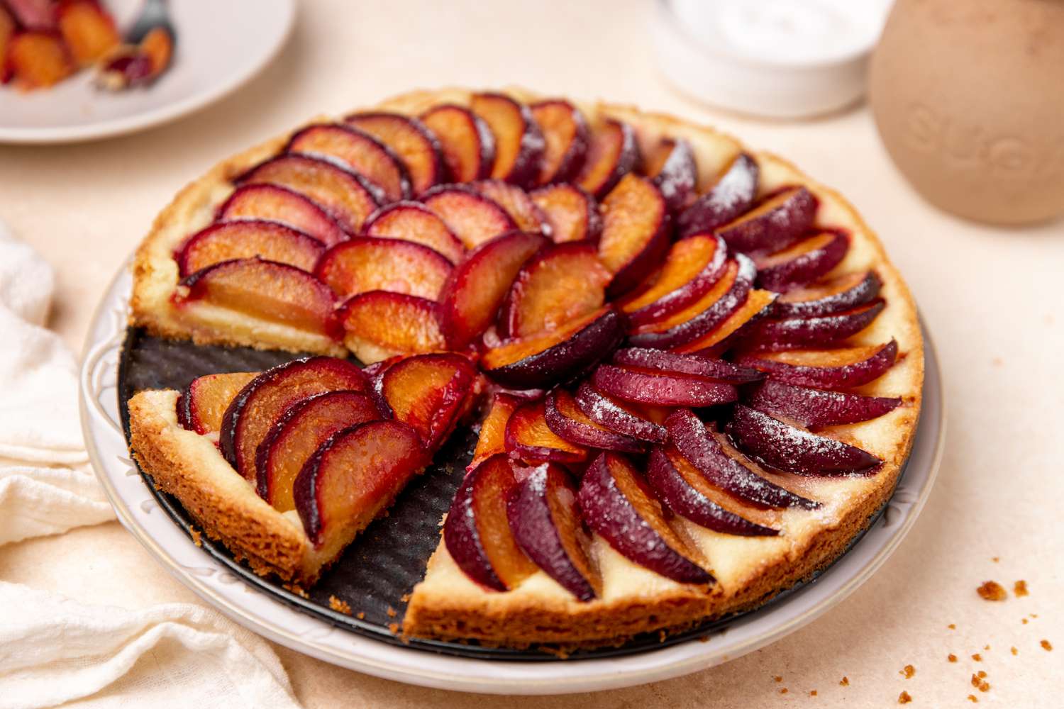 german plum tart (missing a slice) on a plate at a table setting with a kitchen linen, a slice of tart on a plate with a fork, and a bowl of sugar