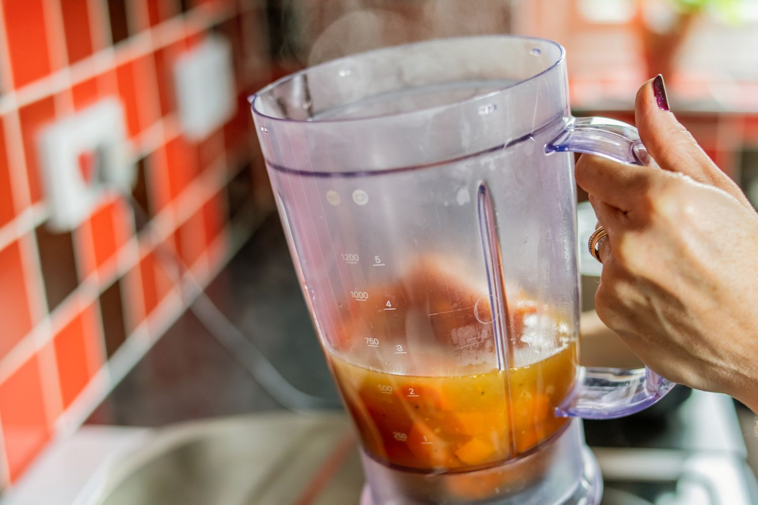 A person holding a blender pitcher filled with hot soup in a kitchen setting with red tiles in the background