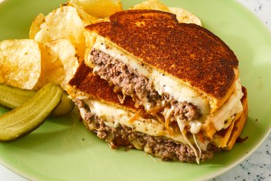 A patty melt sandwich with melted cheese and caramelized onions, served with potato chips and pickle slices on a green plate