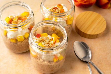 Three jars of peach cobbler overnight oats topped with diced peaches and granola two spoons placed on the table beside them