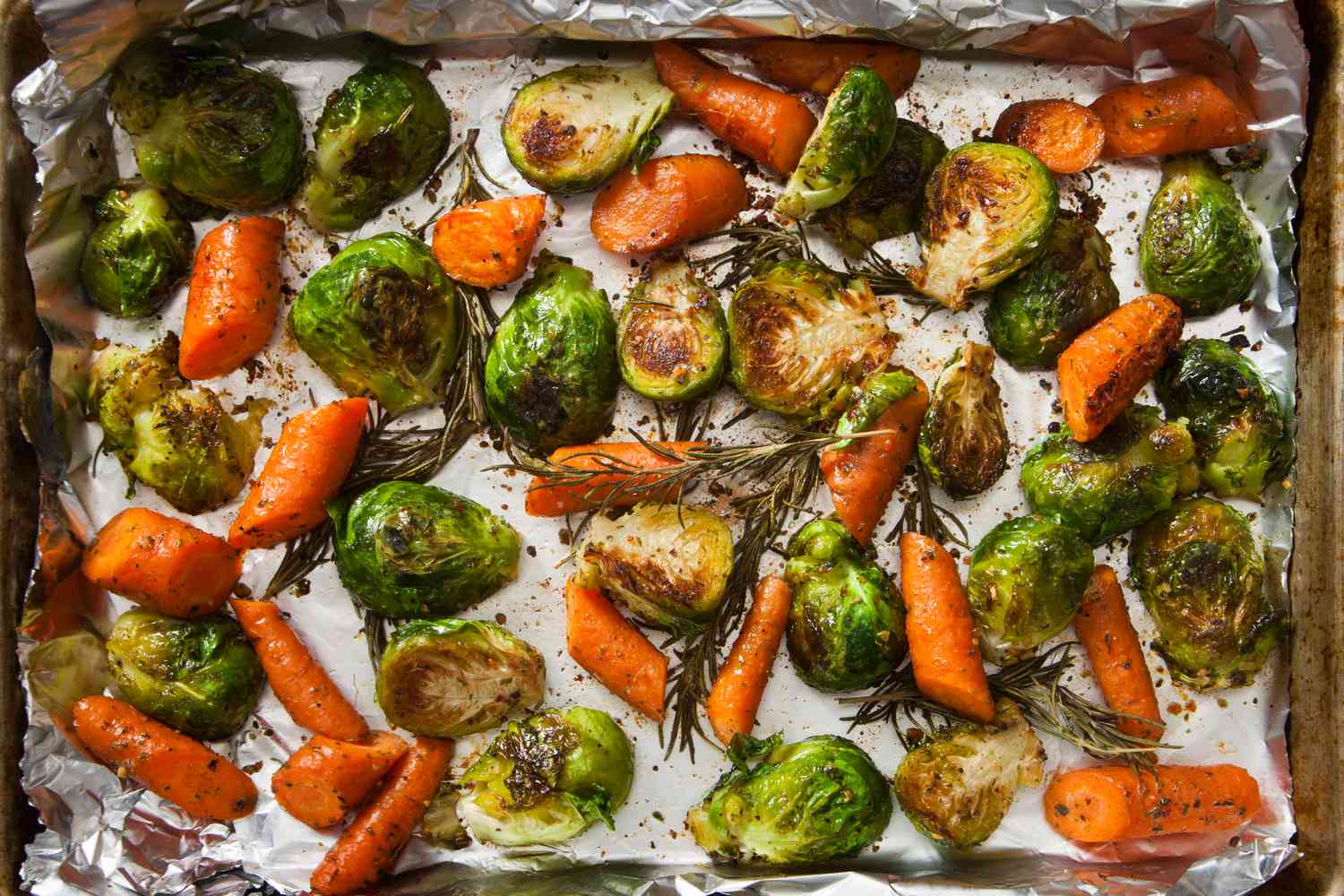 Overhead view of roasted brussels sprouts with rosemary and carrots on a baking sheet lined with foil