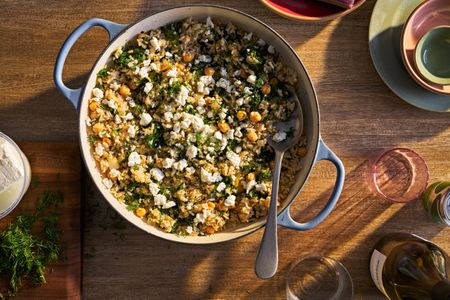 Skillet of chickpea and spinach rice, surrounded by a cutting board of feta and chopped dill, wine glasses, and serving plates