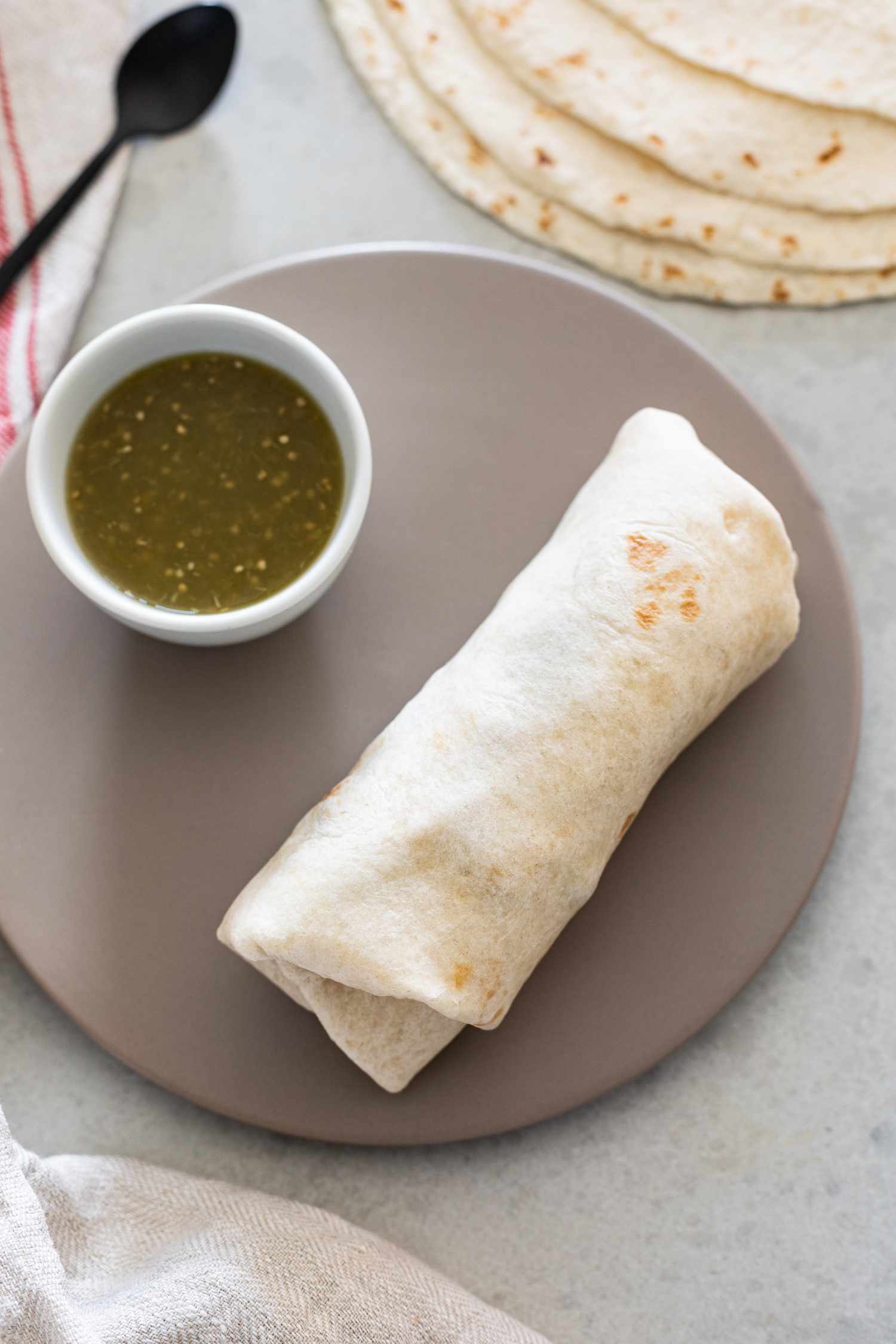 Nicely Folded Burrito on a Plate with a Small Bowl of Salsa and in the Background, a Stack of Tortillas