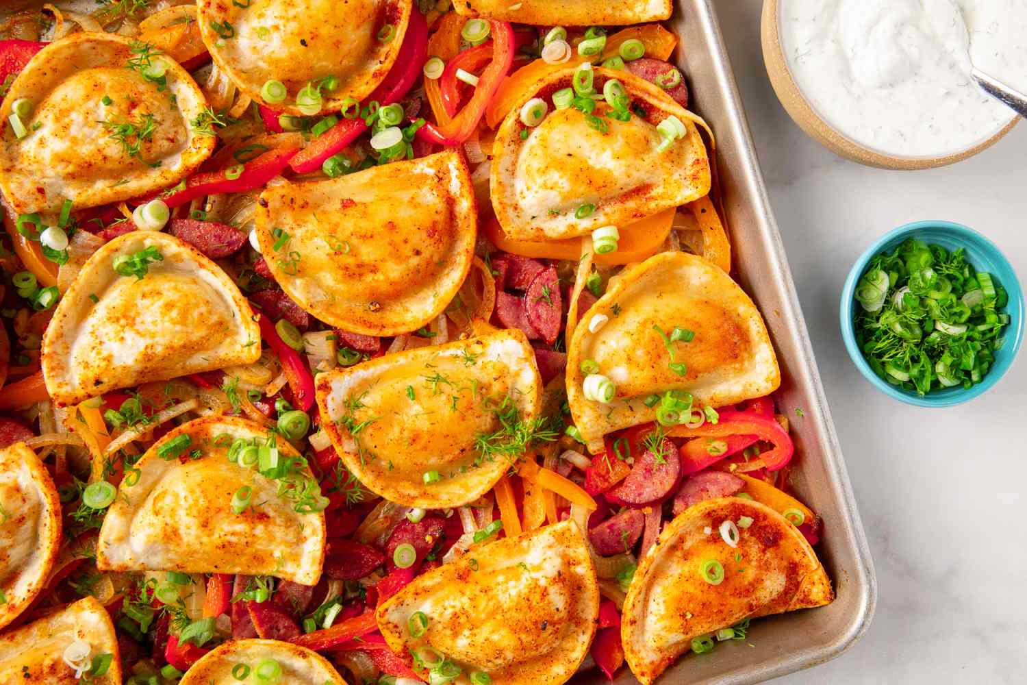 A sheet pan filled with arranged pierogies vegetables and garnished with green onions served next to bowls of sour cream and chopped greens