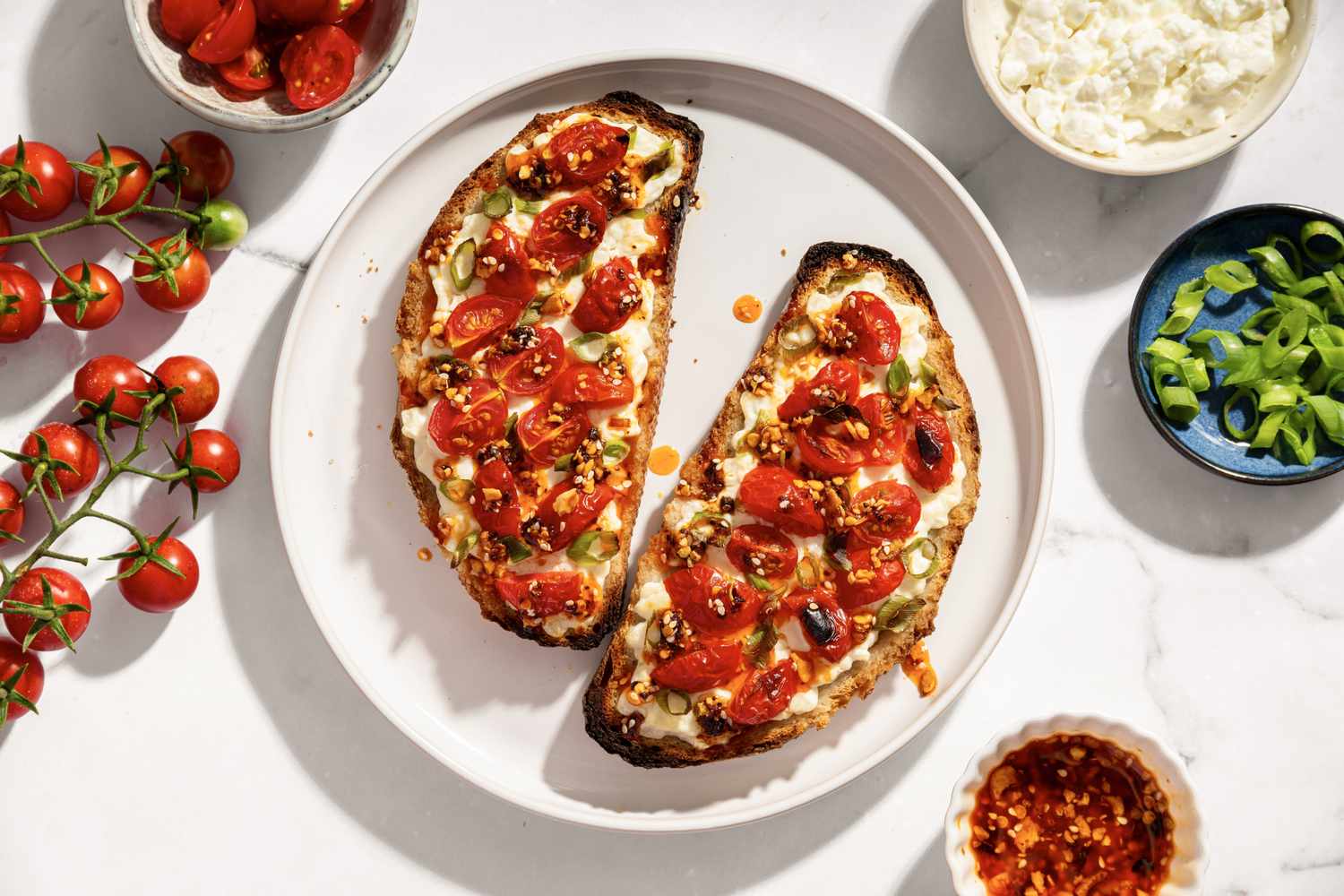 Cottage cheese toast topped with tomatoes and seasonings on a plate surrounded by ingredients like cherry tomatoes and cheese