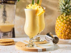 Two Glasses of PiÃ±a Colada With a Pineapple Wedge, Pineapple Leaf, and a Straw on a Marble and Wood Circular Serving Tray, and in the Background, an Uncut Pineapple, a Jigger, and a Blender With Some Leftover PiÃ±a Colada