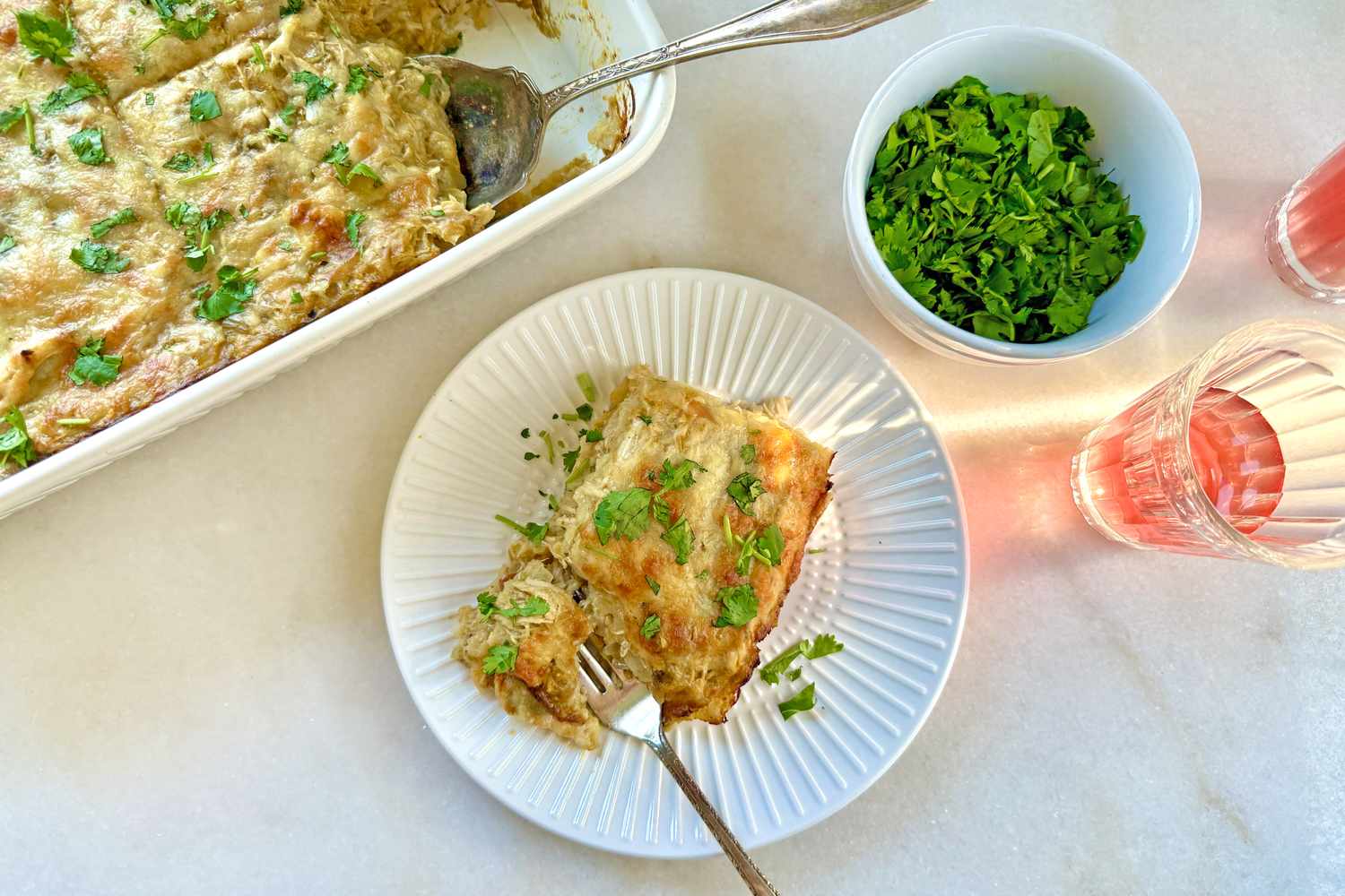 A serving of chicken enchilada casserole on a plate with utensils garnished with herbs with a dish of parsley and glasses nearby