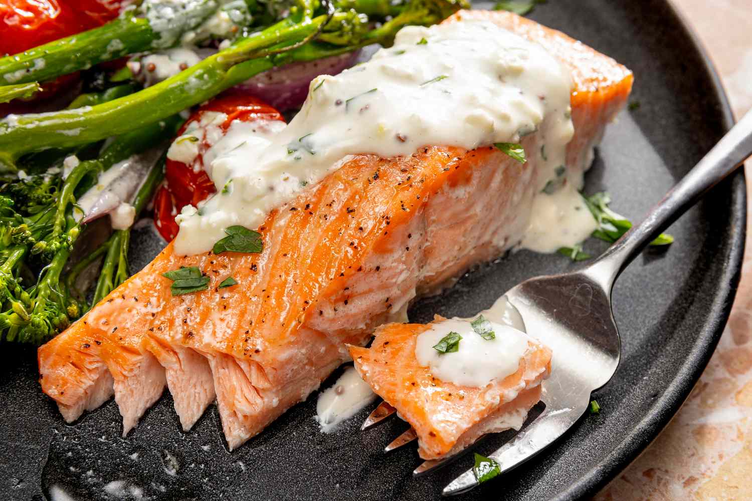 Closeup view of a gray plate of salmon, broccolini cherry tomatoes, onions and feta sauce with a fork on a marble countertop