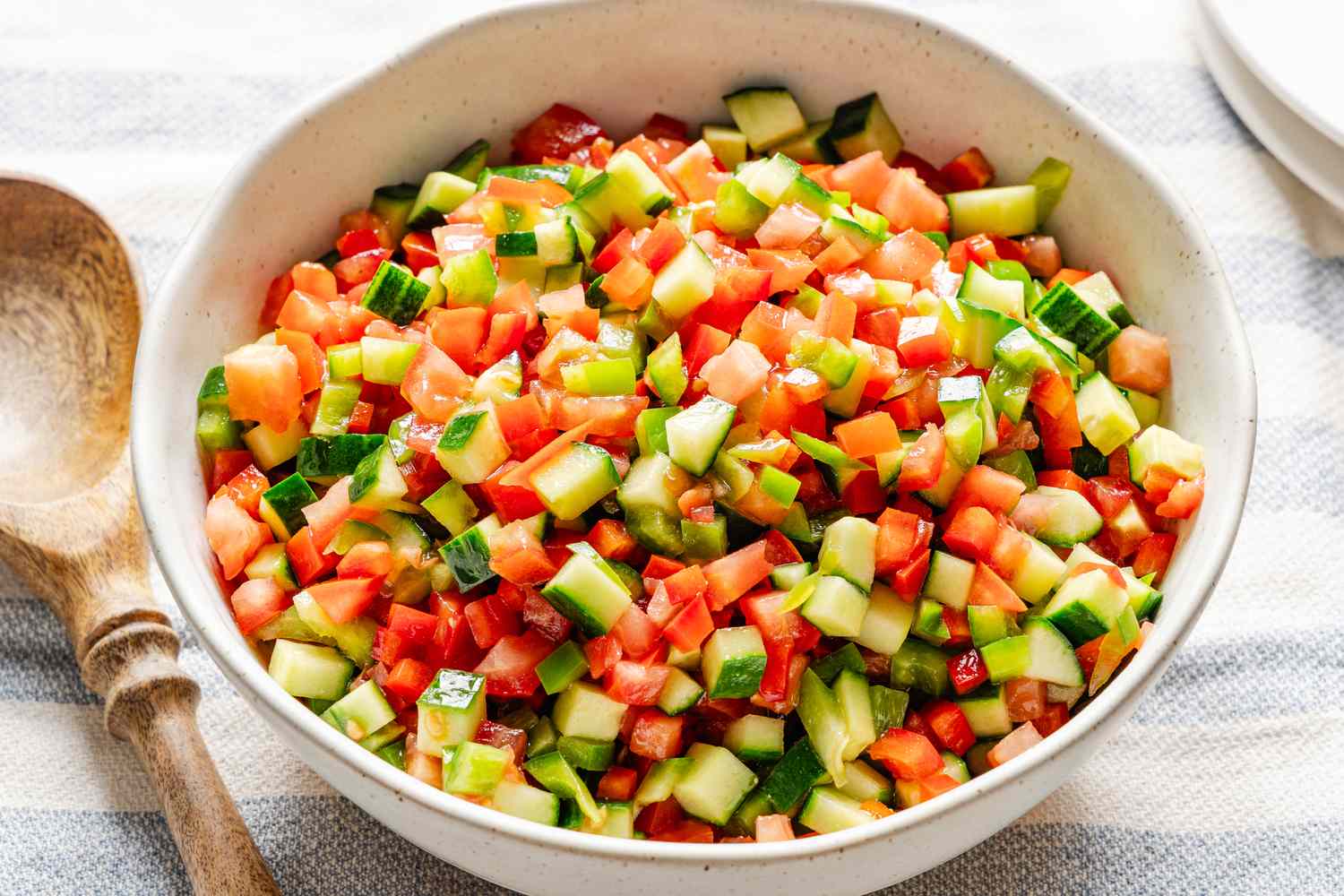 A bowl of diced cucumber tomato and red pepper salad on a table next to a wooden spoon