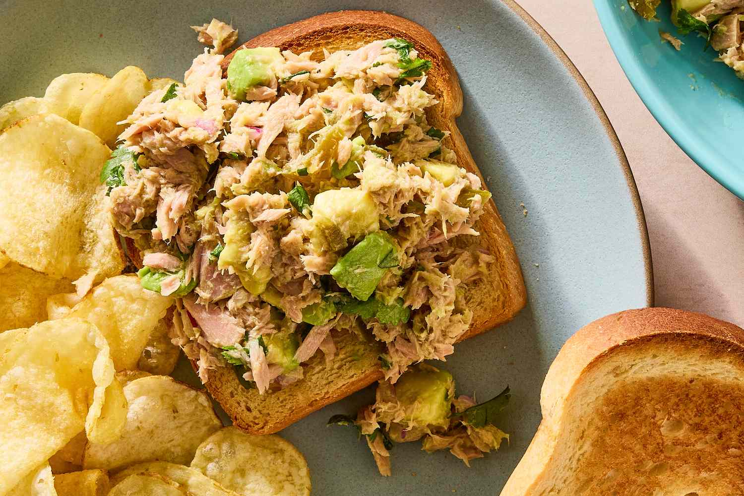 Tuna avocado salad on bread served with potato chips on a plate with additional bread slice