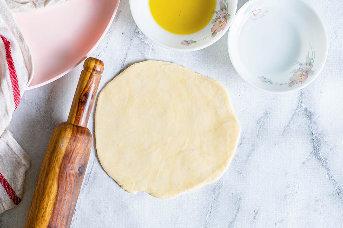 A circle of dough rolled out on a counterop with dishes and a rolling pin nearby.