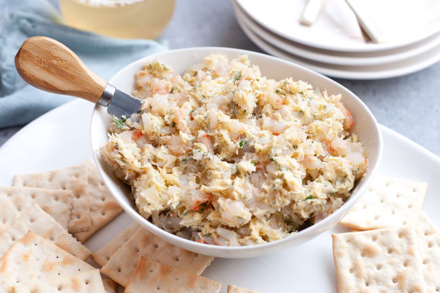 Bowl of Shrimp Butter with a Cheese Knife, Sitting on a Platter of Crackers at a Table Setting with a Glass of Wine, Stack of Plates, and a Kitchen Linen