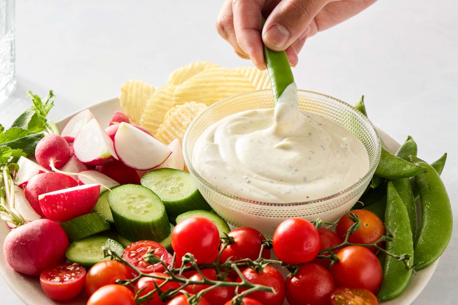 A platter of fresh vegetables and chips with a bowl of ranch dip being used by a hand holding a snap pea
