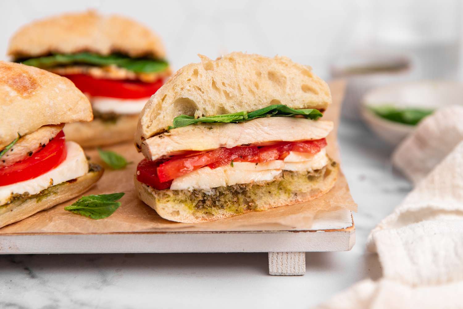 Grilled Chicken Caprese Sandwiches on a Parchment Lined Tray With One Sandwich Cut in Half, and on the Counter Next to the Tray, Plates With Individual Ingredients and a White Kitchen Towel