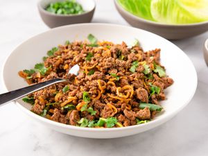 Plate of ground beef larb with sliced herbs, garnished with fried onions, surrounded by small bowls of lettuce and lime wedges