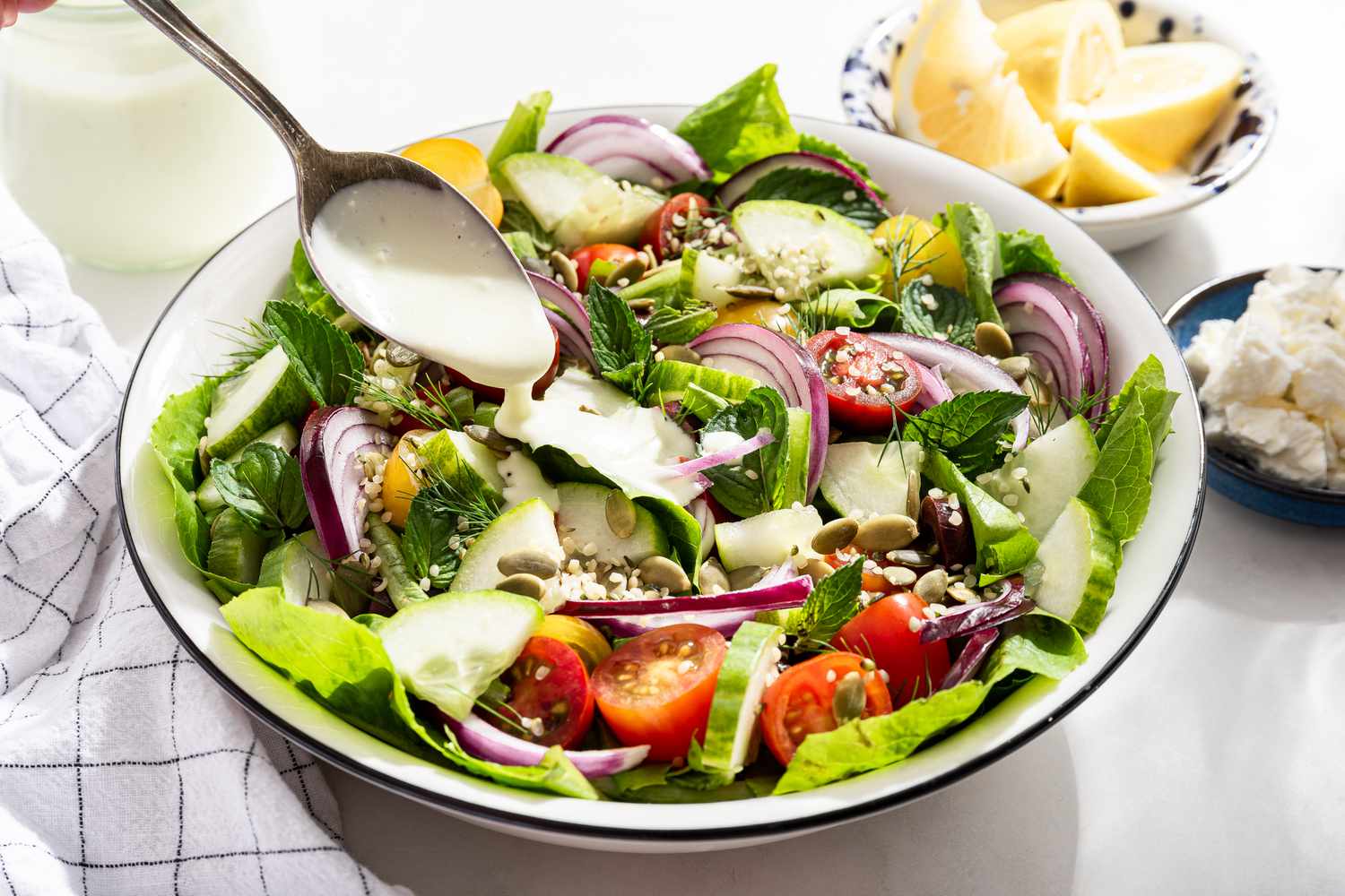 feta dressing spooned onto a salad at a table setting with a bowl of lemon slices, a bowl with feta pieces, a jar with the rest of the dressing, and a table napkin