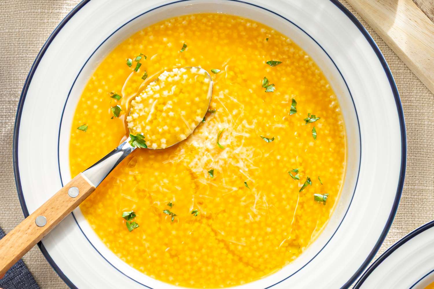 A bowl of pastina in broth garnished with herbs and cheese with a woodenhandled spoon