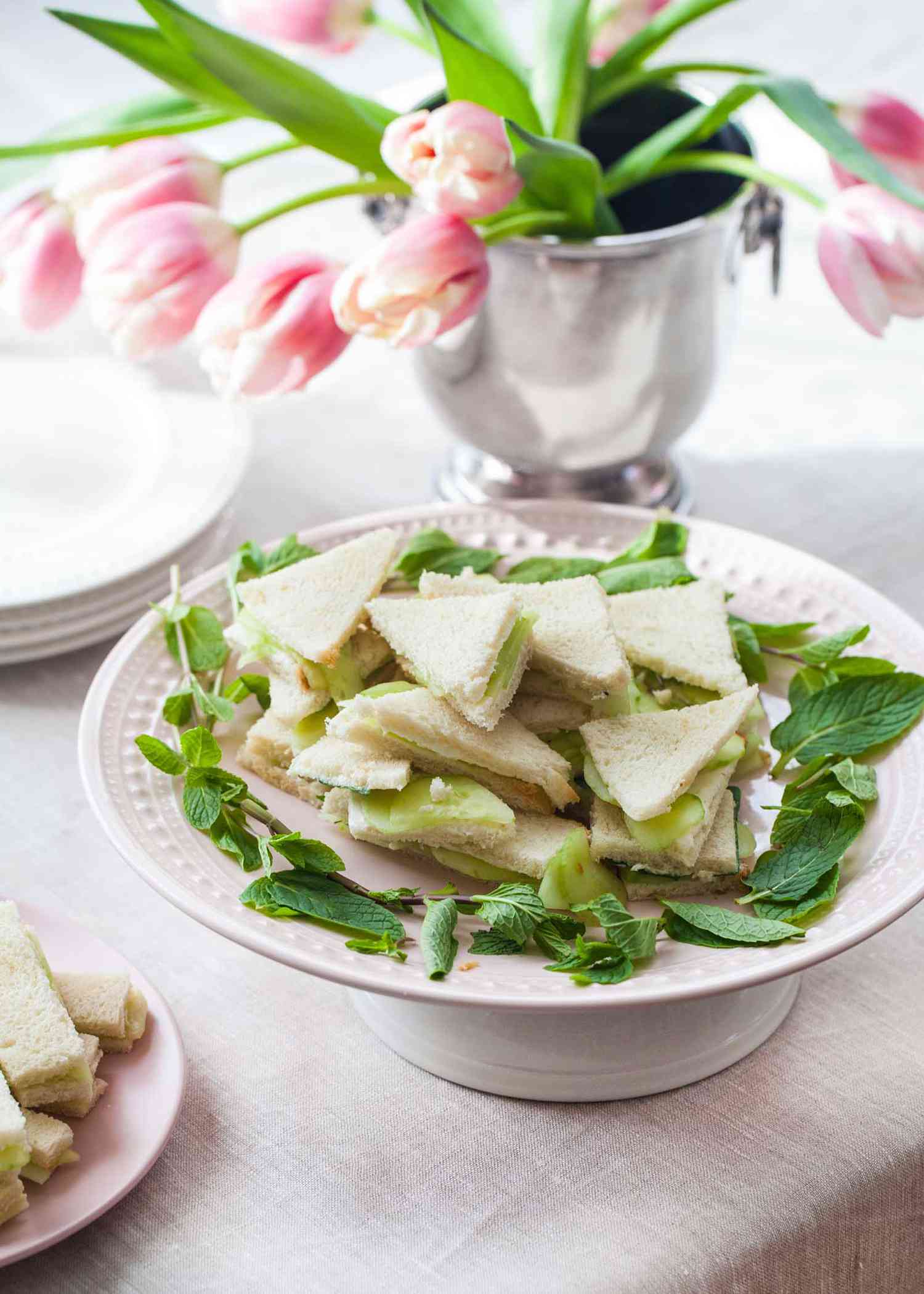 Cucumber cream cheese sandwiches cut in triangles with herbs set around the cake plate they are set on. A boughet of tulips is behind the sandwiches.