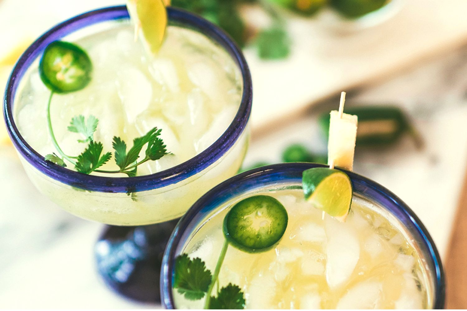 Two margarita glasses, garnished with jalapeño slices, lime wedges, cilantro, and pineapple cubes on skewers, sitting on a table