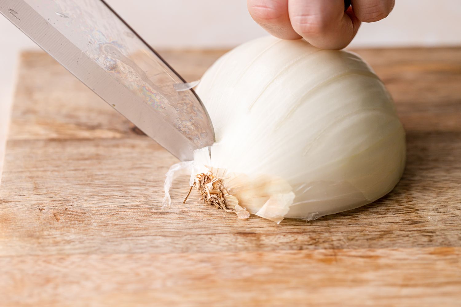 A chef's knife slicing the root end off of a white onion on a cutting board 