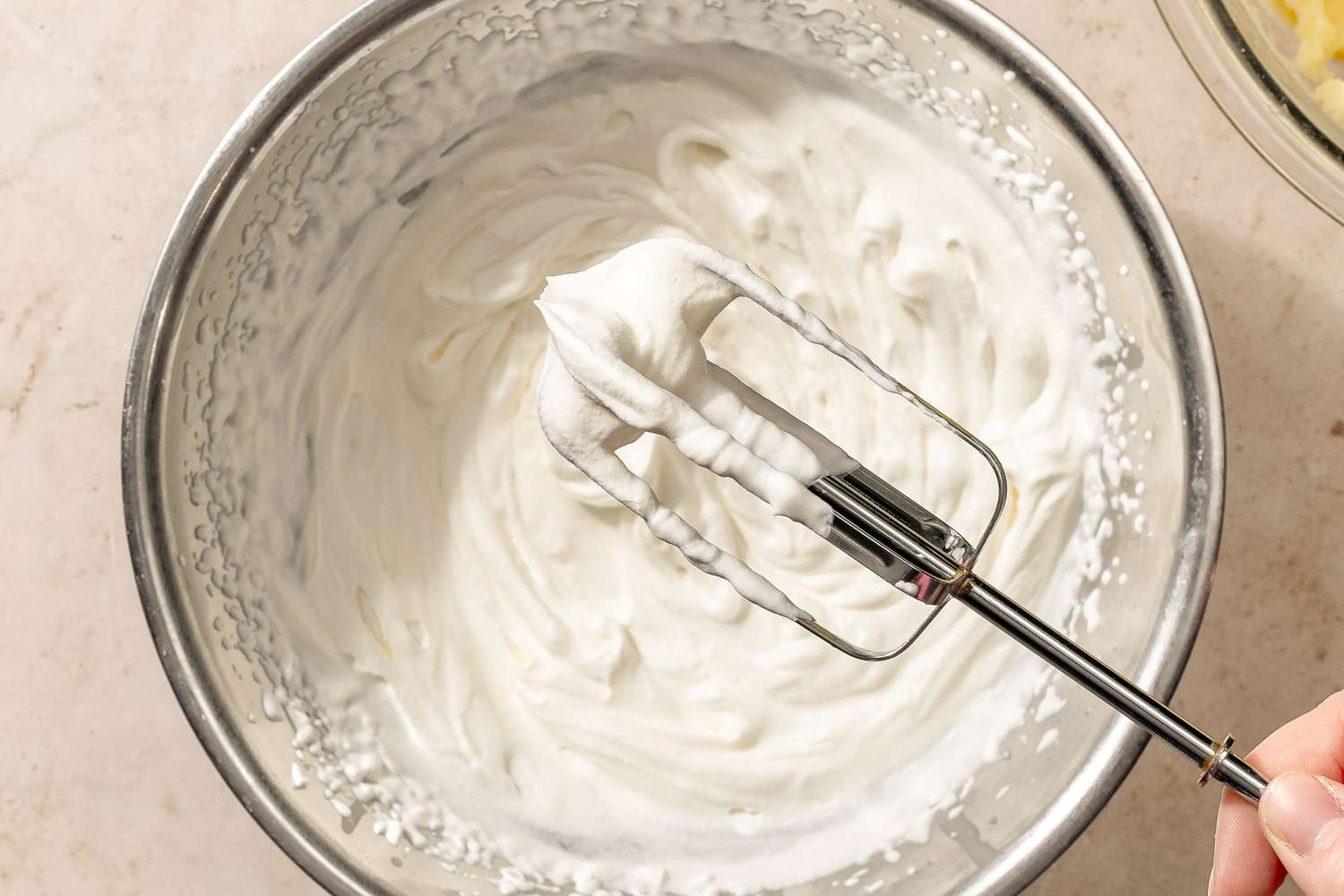 overhead view of whipped cream in a mixing bowl with a hand holding a mixer attachment for Chantilly Potatoes recipe