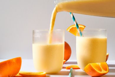 Two glasses of orange smoothie are served, with a blue-striped straw in one and orange slices on the rim and table