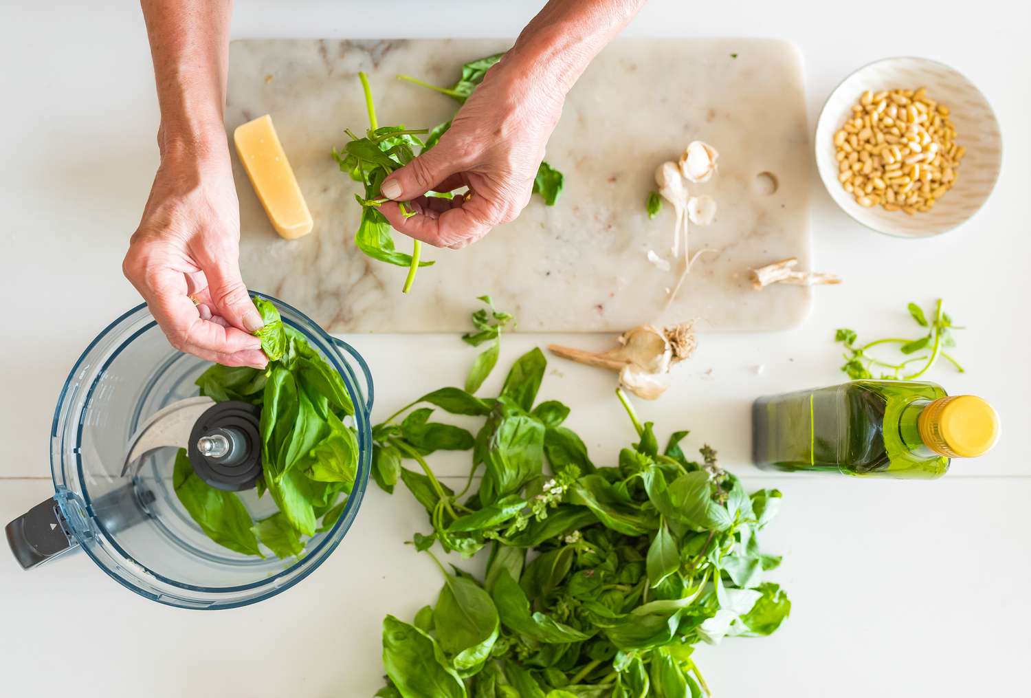 Hands adding basil leaves to a mini food processor with ingredients for pesto