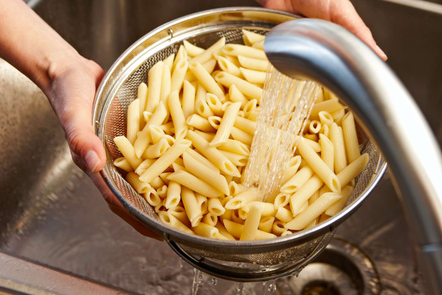 Person rinsing penne pasta in a colander under running water in a kitchen sink